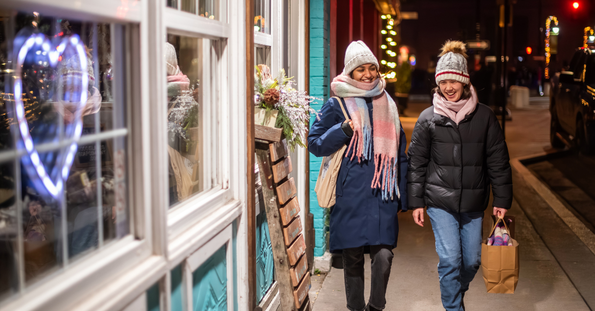 Two women walking outdoors with holiday decorations and a heart-shaped light.
