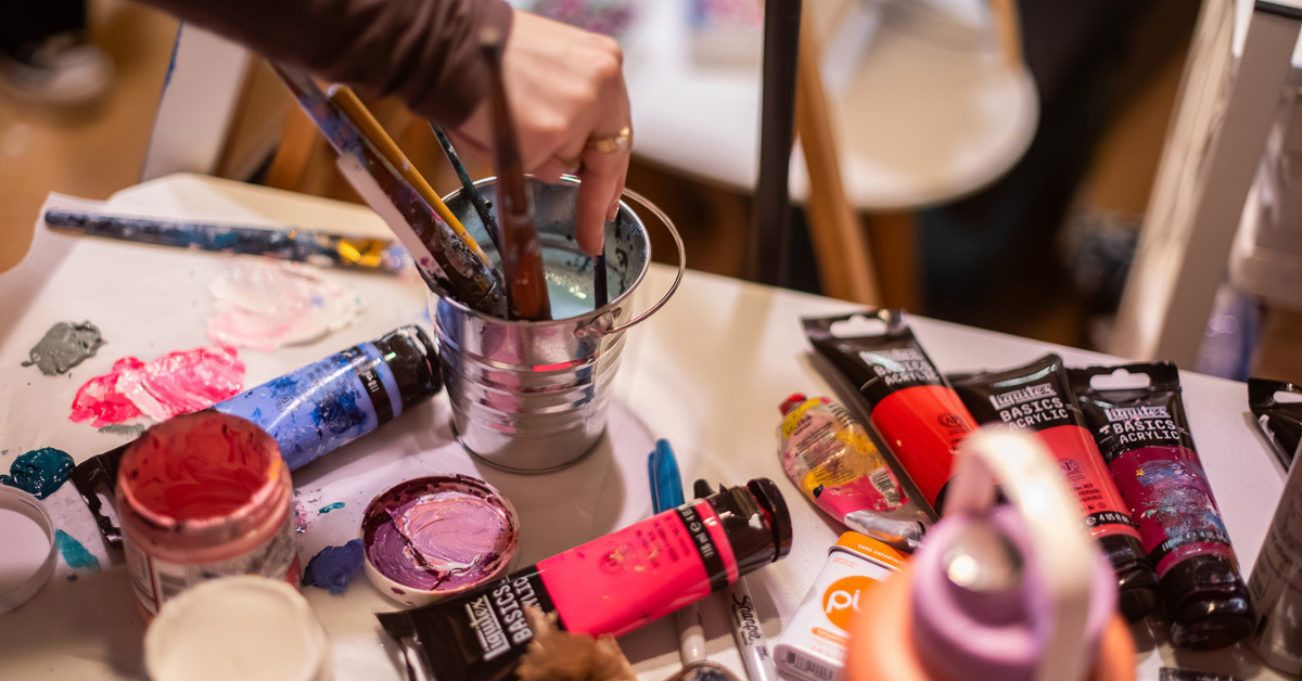 A person's hand holding a brush over paint supplies on a table.