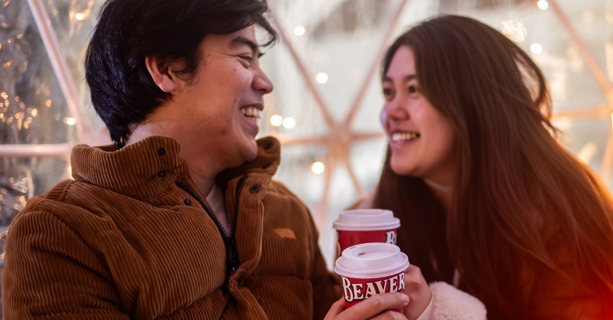 A couple smiling and holding coffee cups in a cozy setting.
