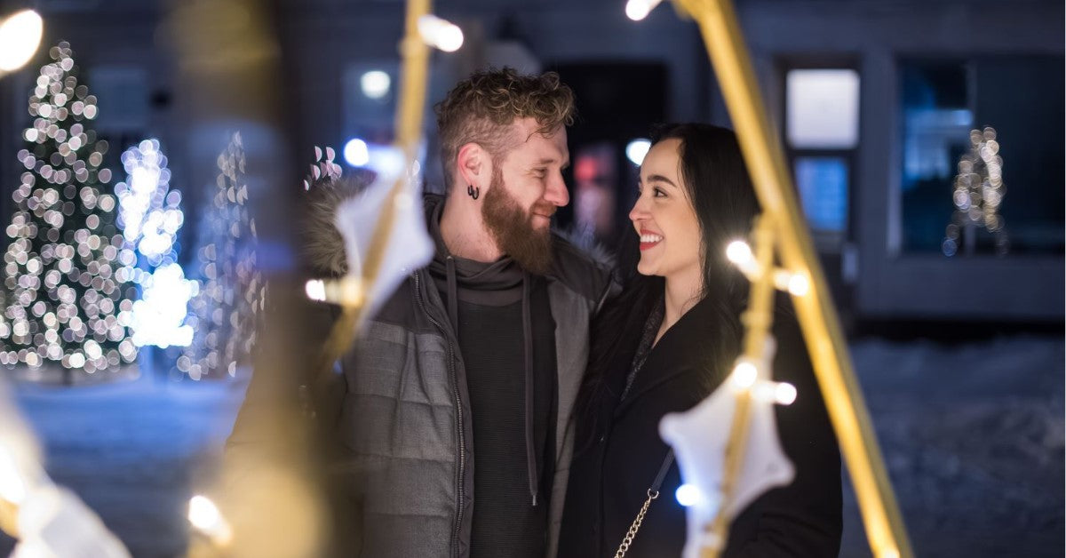 Couple smiling at each other surrounded by festive lights and decorations.