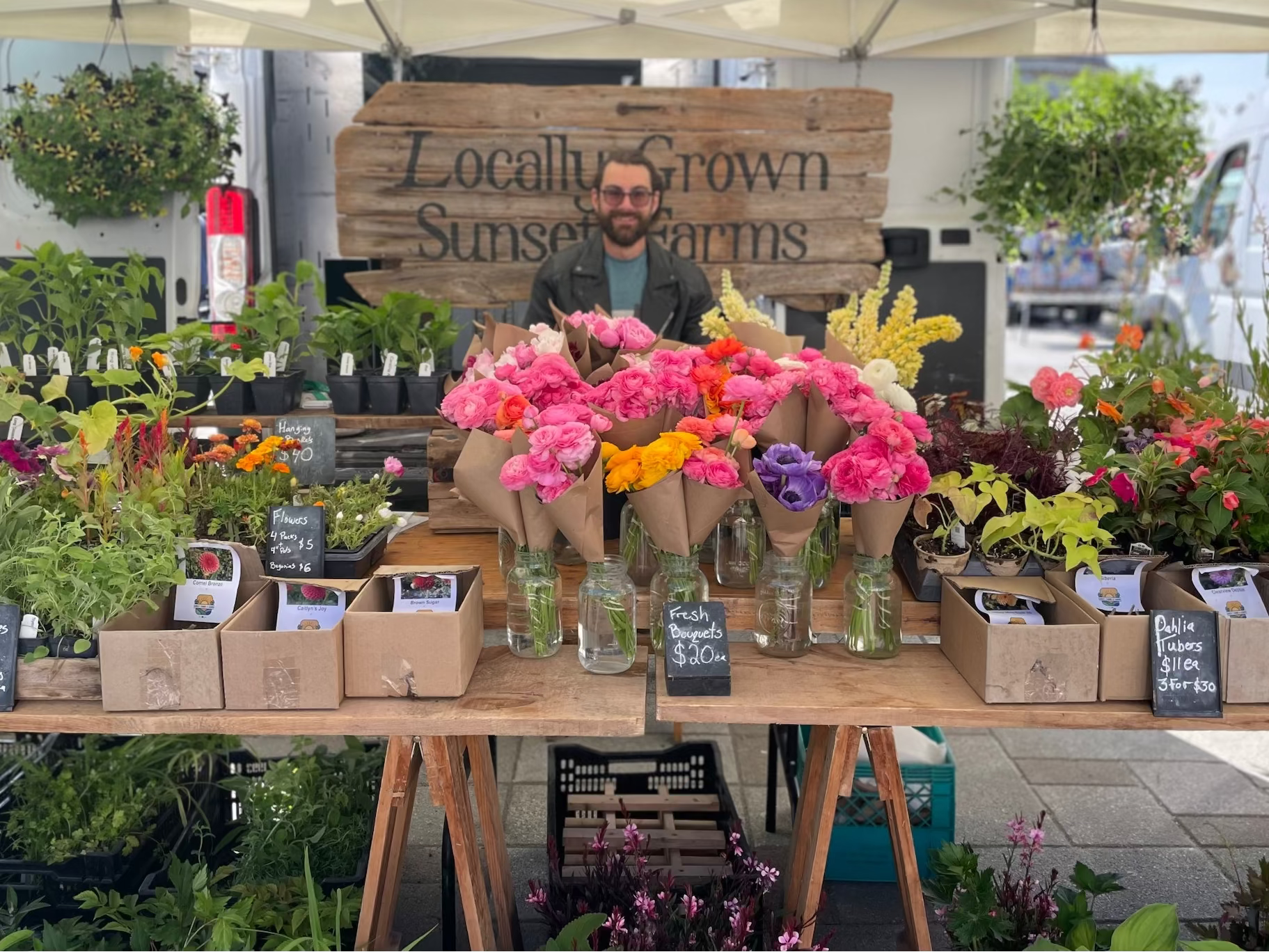 Man standing behind a table of flowers and plants at a market stall with 'Locally Grown Sunset Farms' sign.