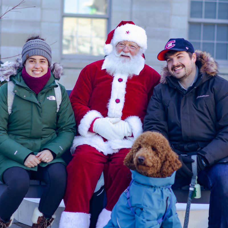 Person in Santa Claus costume sitting with two people and a dog outdoors.