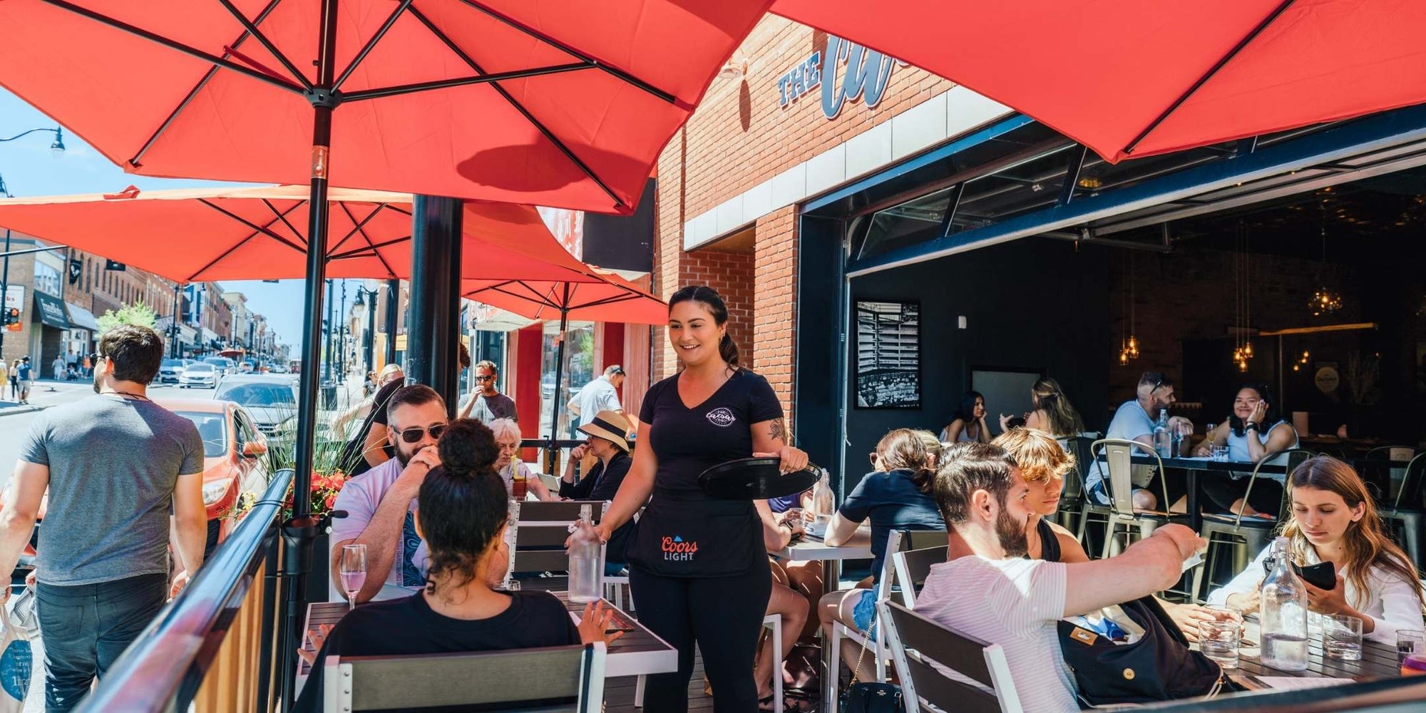Busy outdoor restaurant patio with diners under red umbrellas in downtown Kingston.
