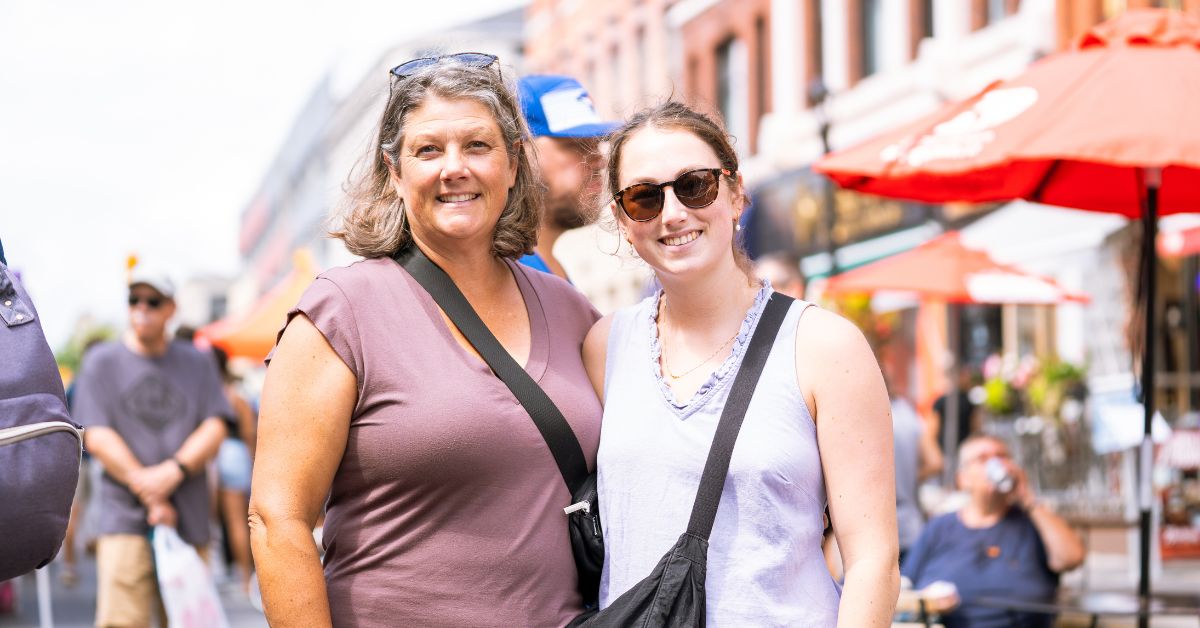 Two women smiling together in a lively outdoor setting.