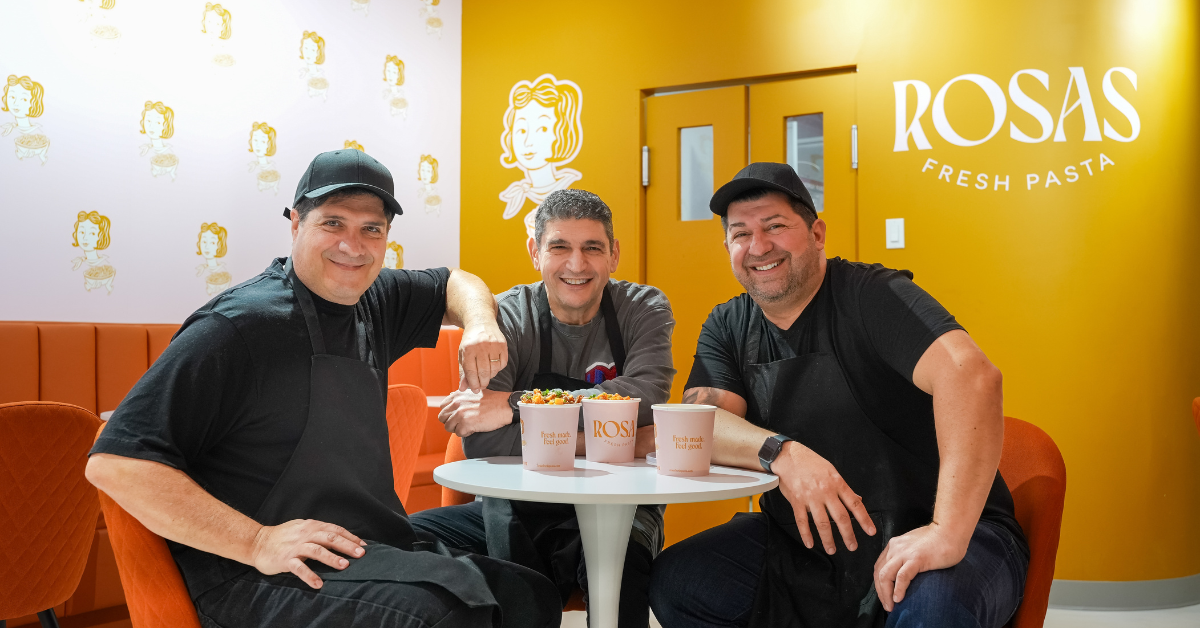 Three men smiling at a table with bowls of pasta in a colorful restaurant.