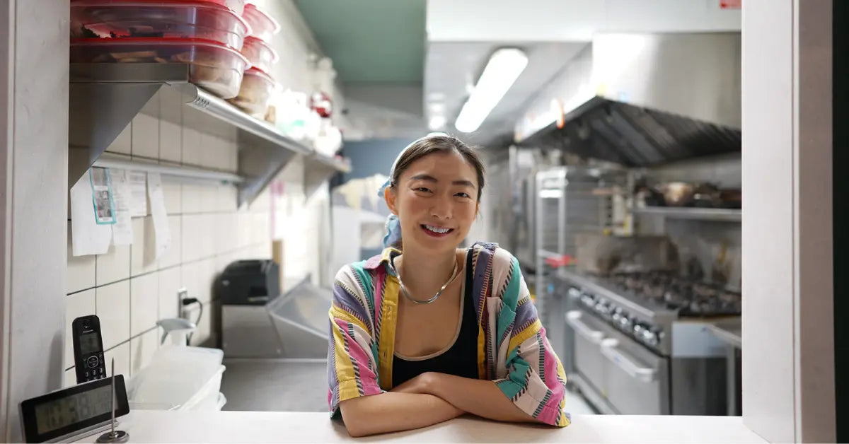 Bellen Tong smiling in Miss Bao's kitchen, wearing a colorful shirt, with cooking equipment visible.