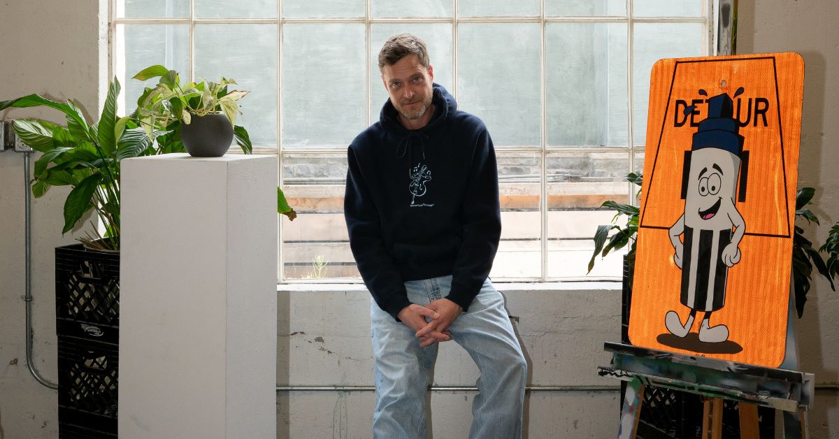 Person sitting beside an orange painting and a potted plant indoors.