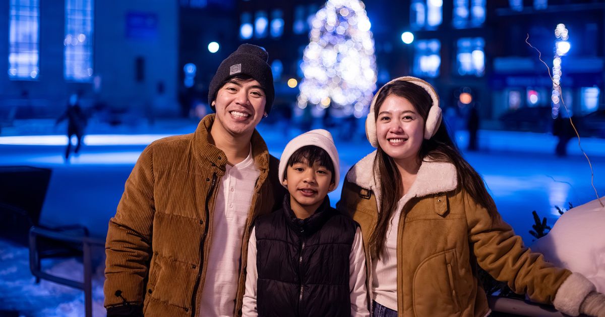 A family of three, a dad, mom and son, smiling together outdoors near a holiday tree at night.