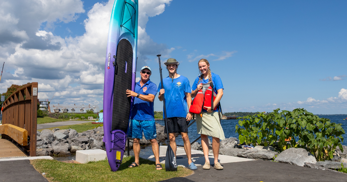 Three people with kayaks by the water on a sunny day.