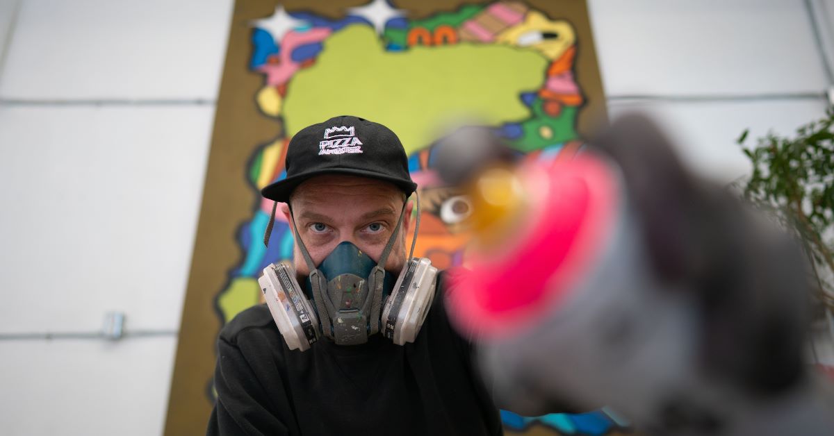 Person wearing a mask, holding a spray paint can in front of colorful background.