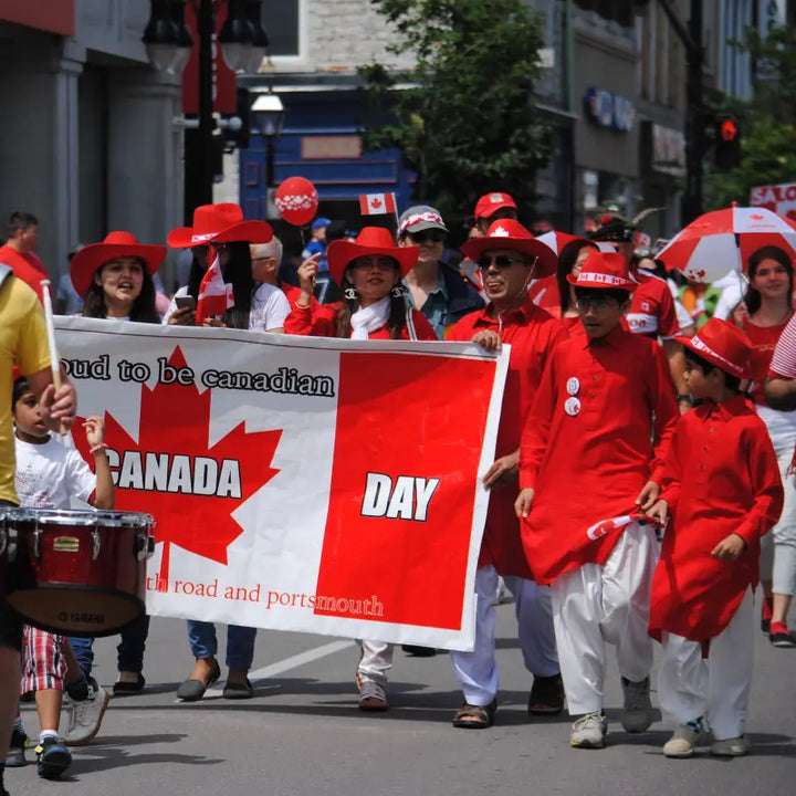 A group of people dressed in red and white walk in a Canada Day parade, proudly holding a large banner with a red maple leaf that reads "Proud to be Canadian – Canada Day." Many participants wear red cowboy hats, wave Canadian flags, and smile as they move down a city street filled with spectators and festive decorations.