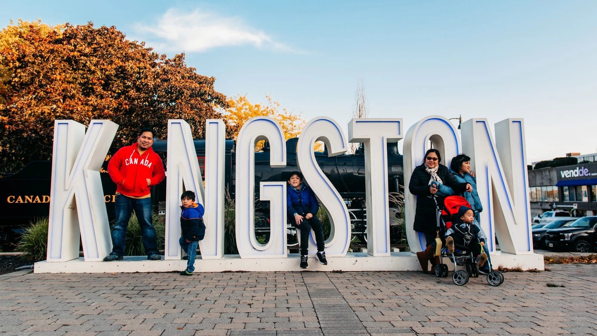People posing in front of a large 'Kingston' sign with trees and buildings in the background.