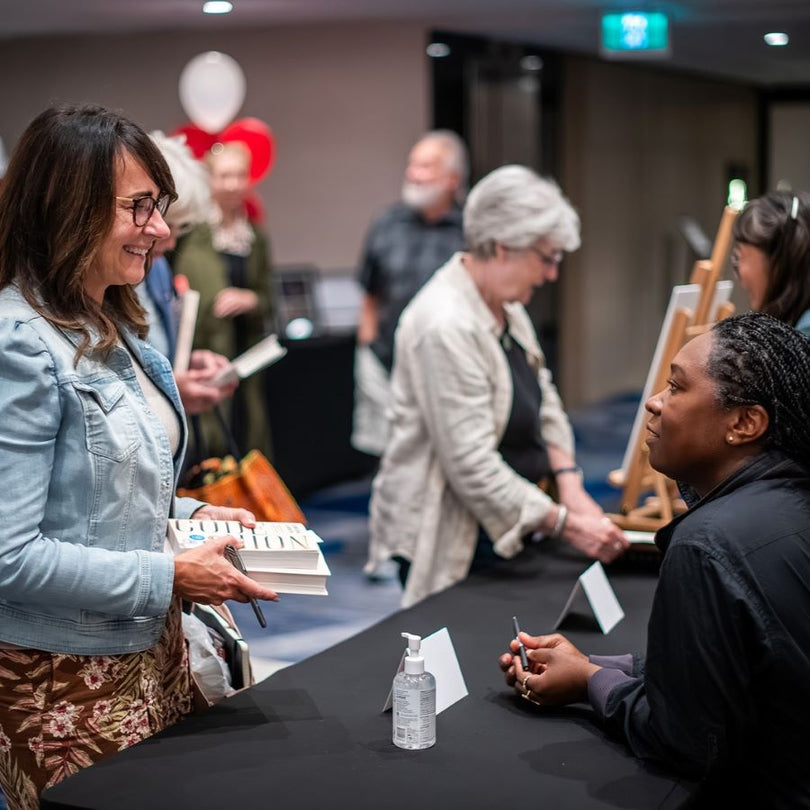 A woman in a light blue denim jacket smiles while holding books at a table, with other people and an artist easel visible in the background of this indoor event scene.