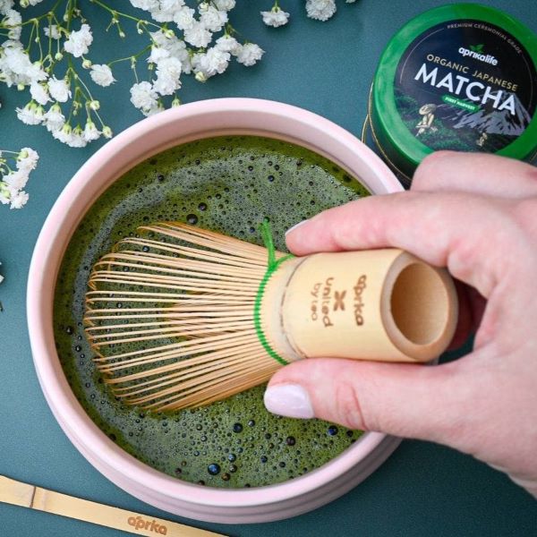 Person using a wooden matcha whisk to stir matcha green tea in a bowl with a container of aprilla matcha in the background.