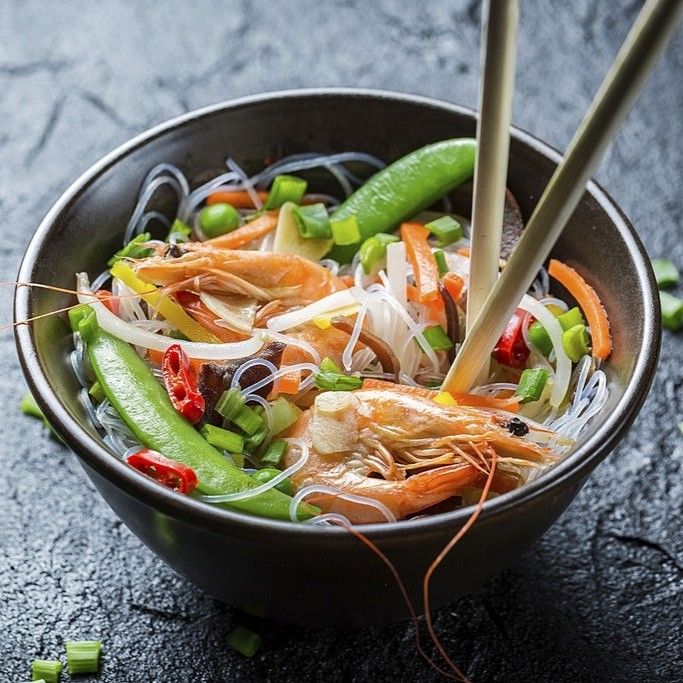 Bowl of noodle salad with shrimp, vegetables, and chopsticks on a dark surface