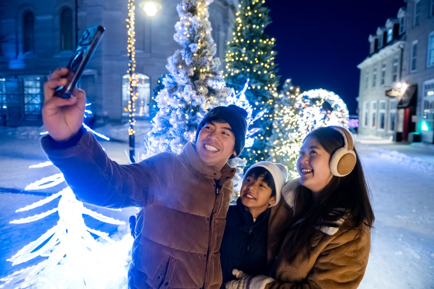 Three people taking a selfie in front of illuminated trees at night.