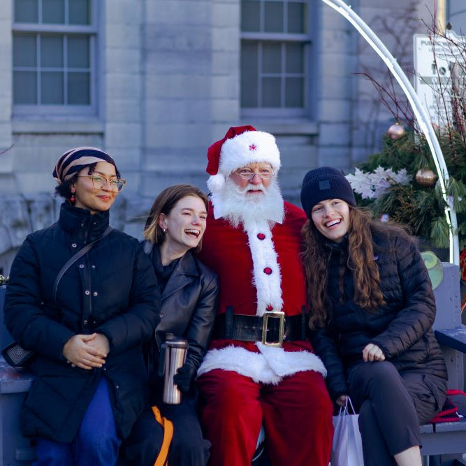 Santa Claus sitting with three women on a bench outdoors, with Christmas decorations in the background.