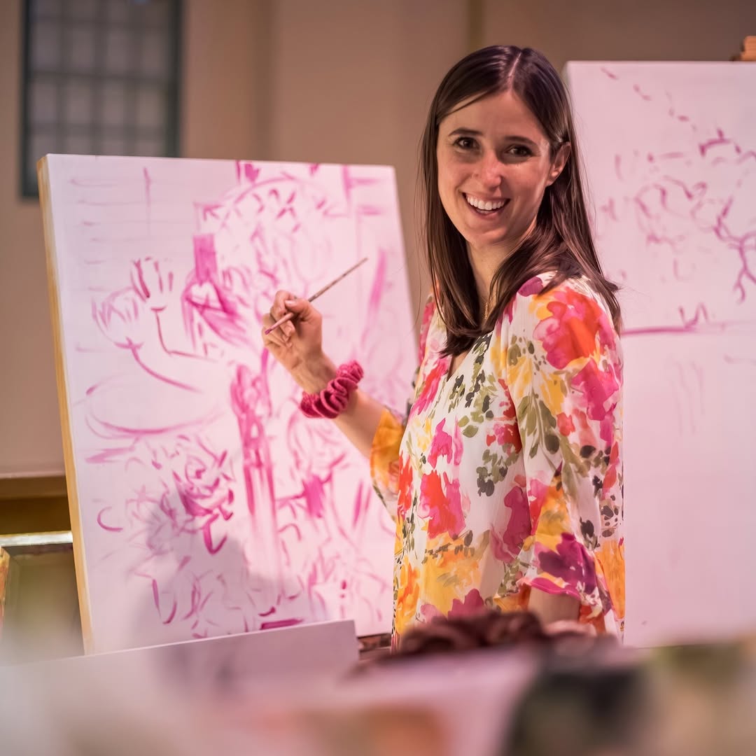 Woman in a floral shirt painting on a canvas with pink abstract design.