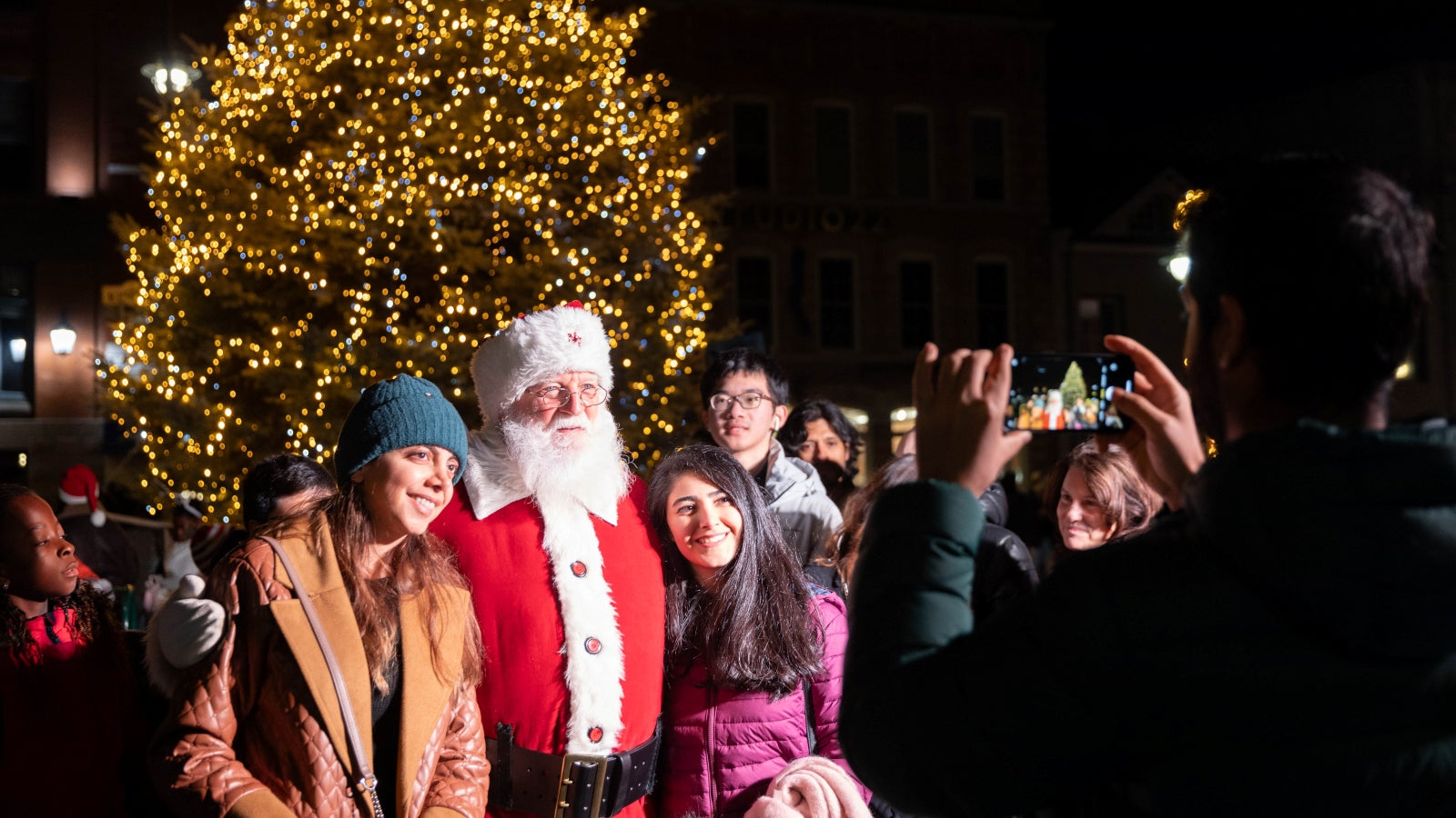 People taking photos with Santa Claus in front of a Christmas tree at night.